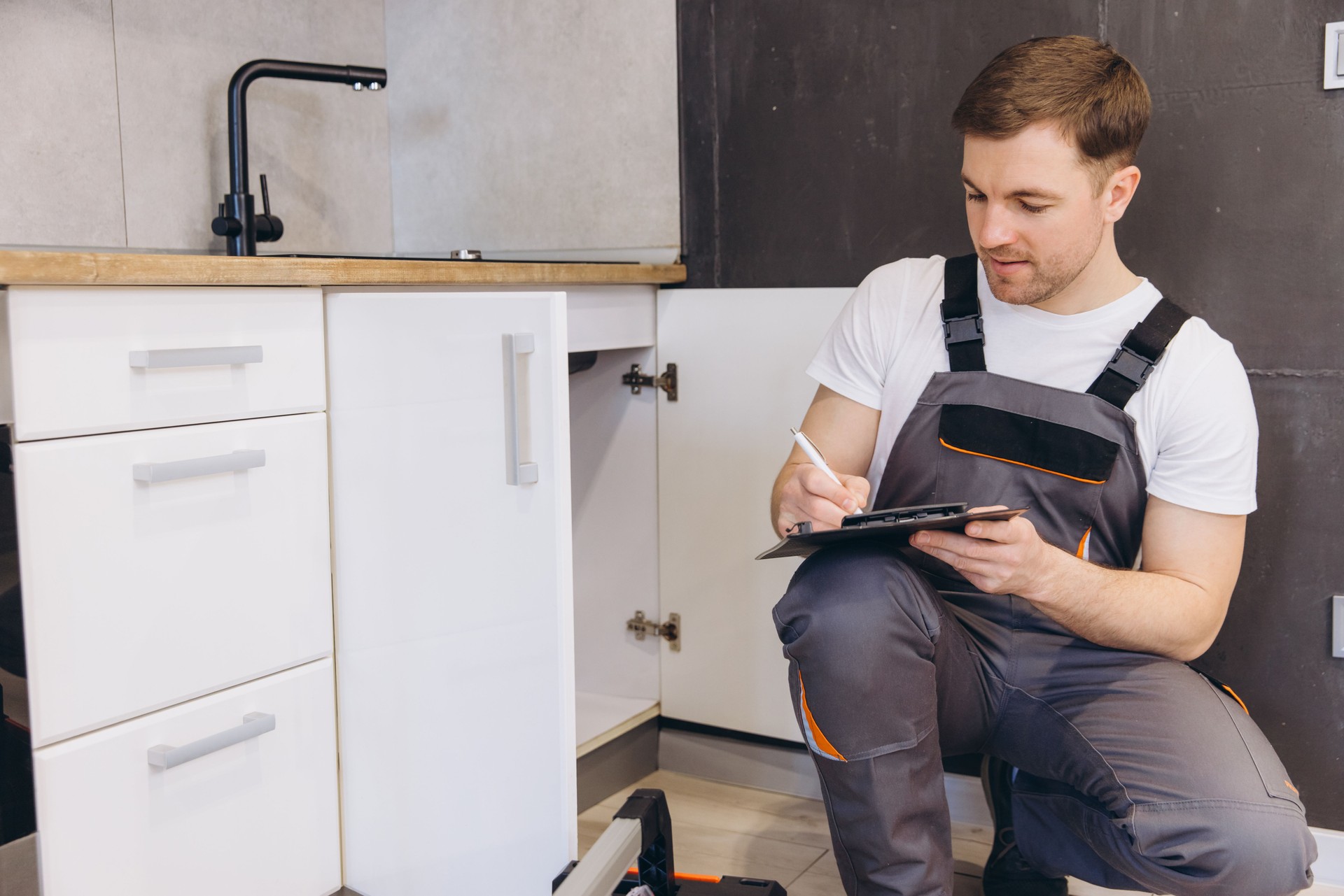 Plumber writing on clipboard inspecting pipes under kitchen sink